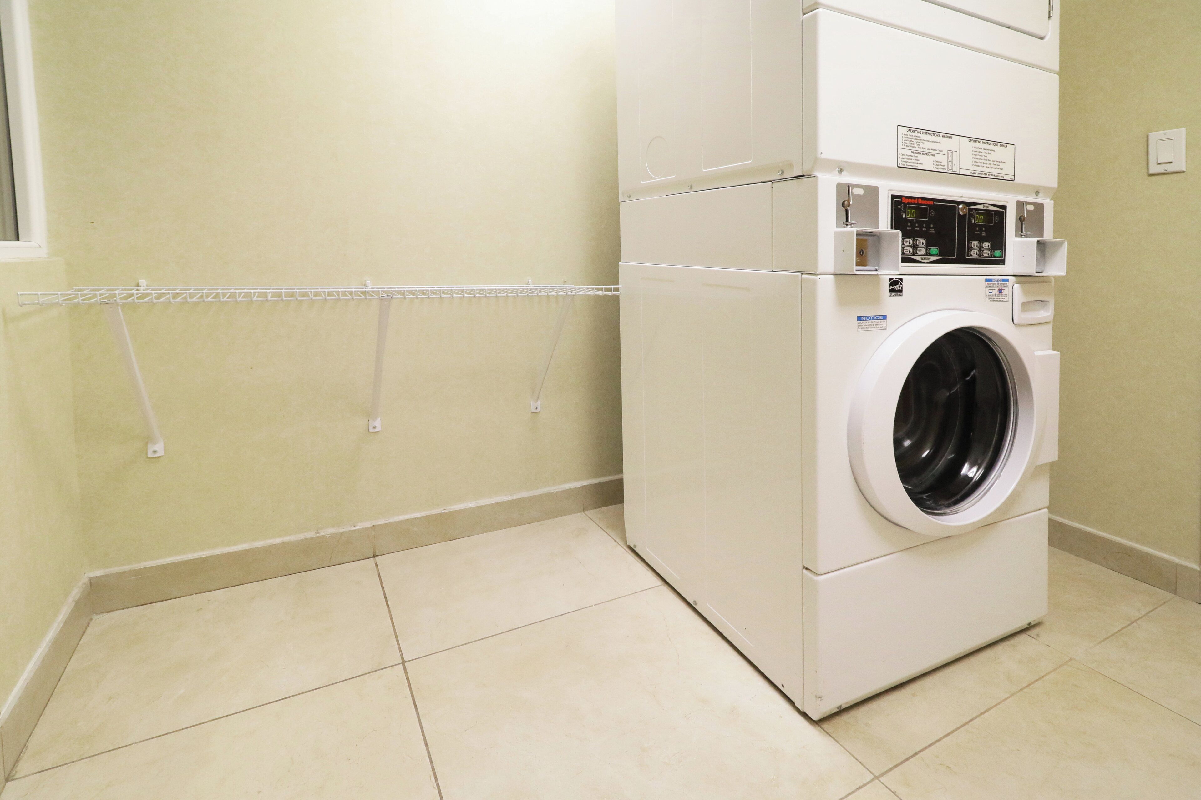A compact, white, wall-mounted washer and dryer unit with a digital display and control panel, installed in a corner of a room with plain walls and tiled flooring.