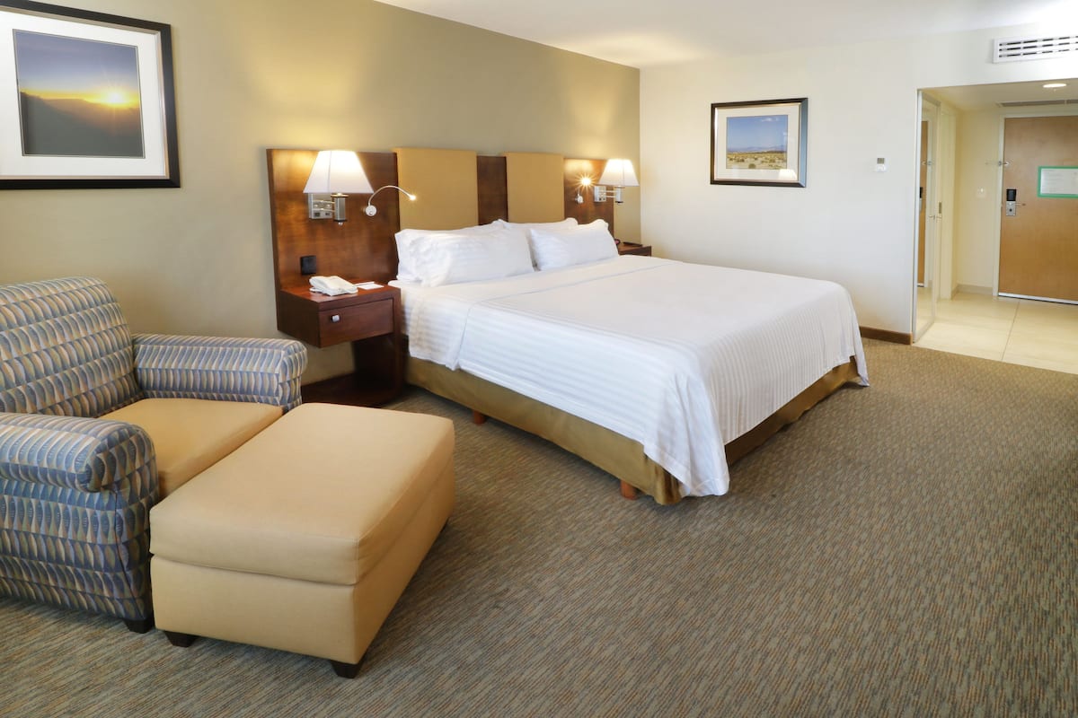 A hotel room with a bed, a chair, an ottoman, a nightstand, and two wall-mounted lamps.