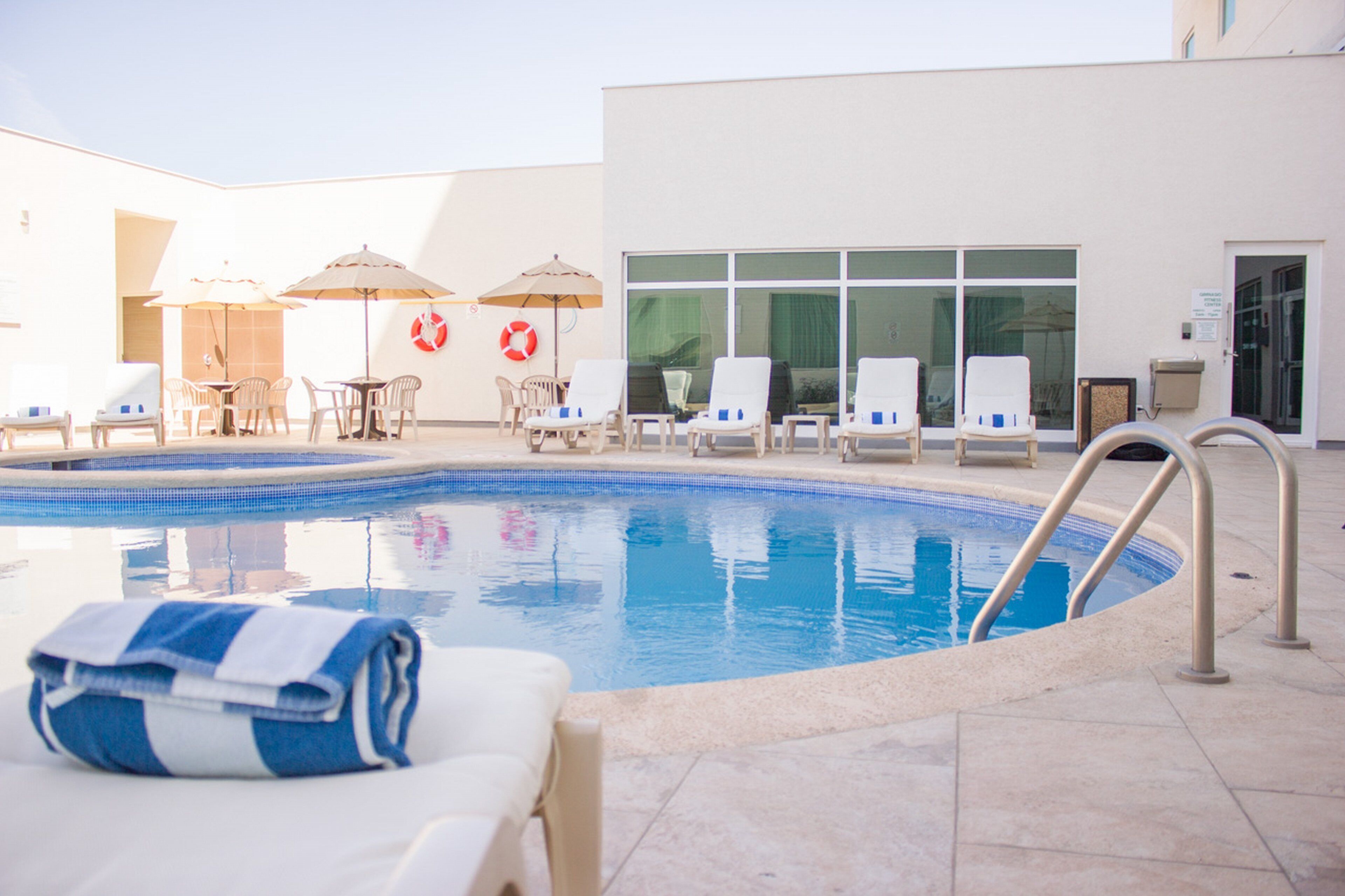 A swimming pool with lounge chairs and umbrellas, a lifebuoy, and a building entrance.