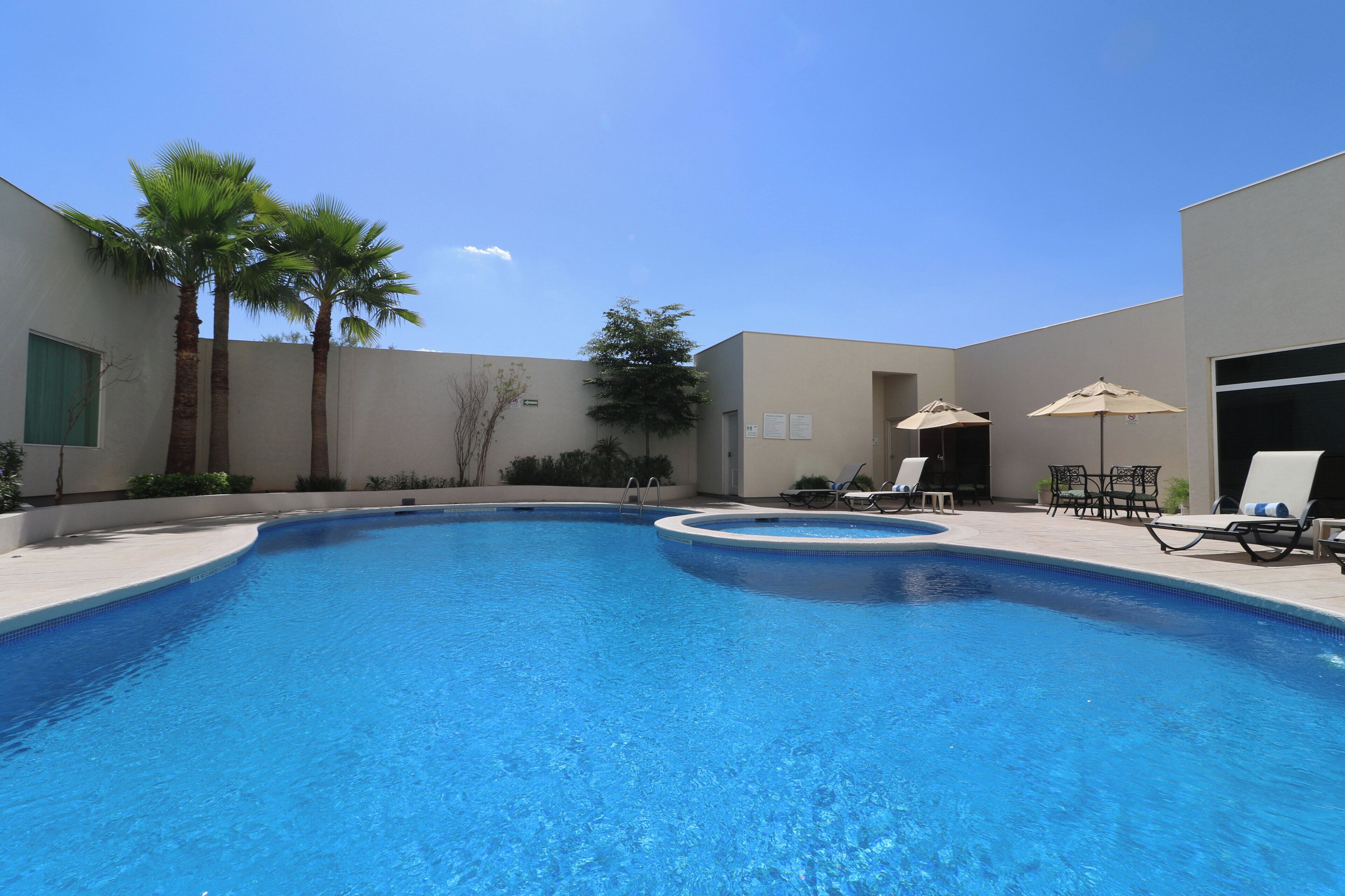 A swimming pool with lounge chairs and umbrellas, surrounded by palm trees and a building.