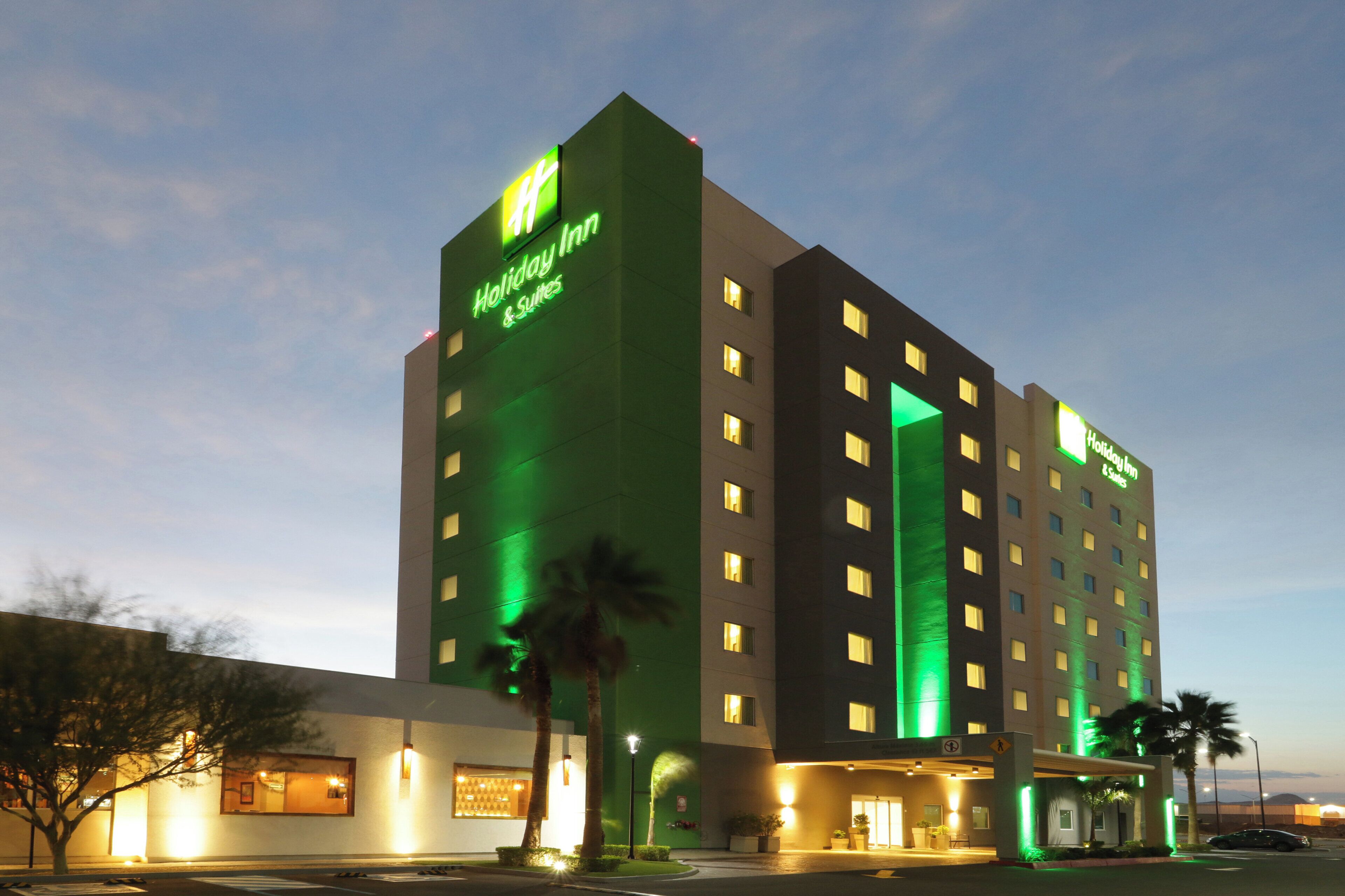 A multi-story Holiday Inn hotel with a prominent green neon sign, multiple windows, and a parking area with palm trees.