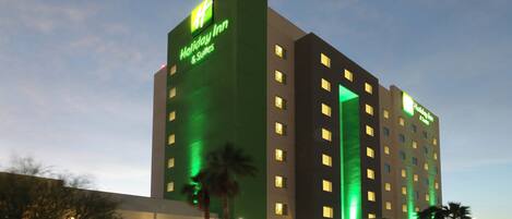 Un hotel Holiday Inn de varios pisos con un llamativo letrero de neón verde, numerosas ventanas y un estacionamiento con palmeras.