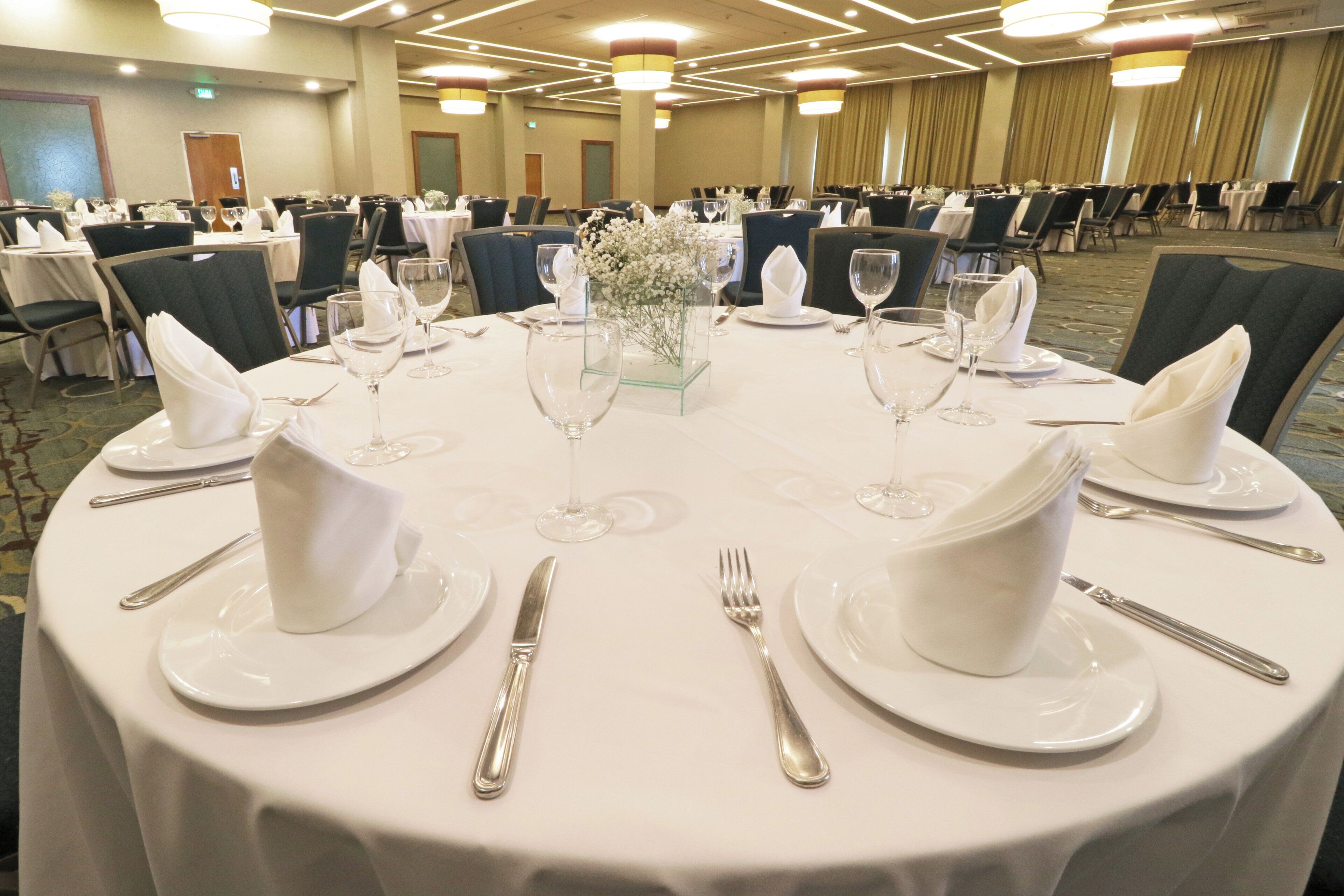 A banquet hall with round tables set for dining, featuring white tablecloths, plates, cutlery, and glassware.
