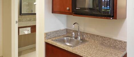 Una cocina con encimeras de granito, un lavamanos de acero inoxidable, microondas y armarios de madera.