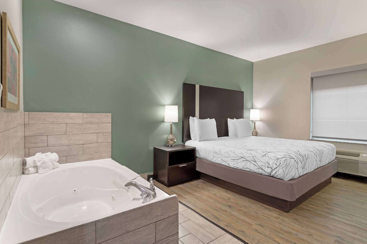 A modern hotel room with a large bed, a separate bathroom with a bathtub, and a nightstand with lamps.