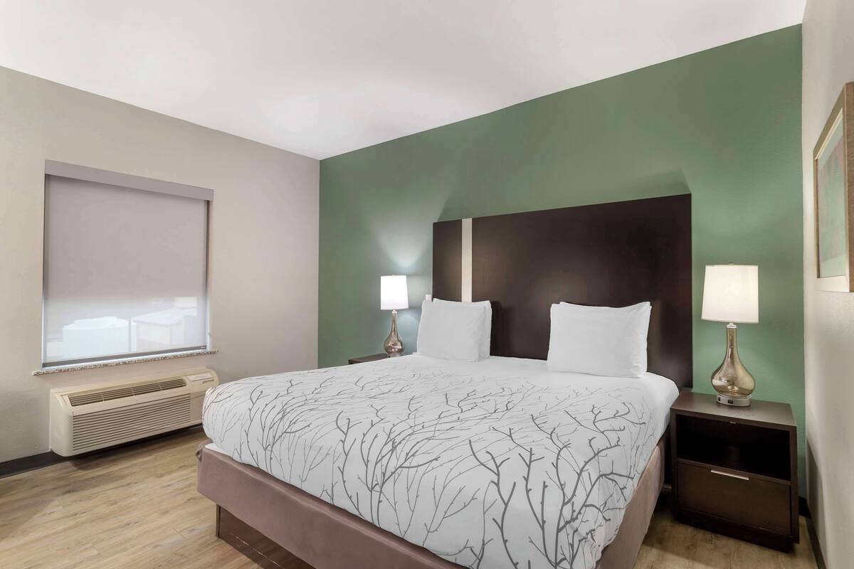 A hotel room with a large bed, two bedside tables with lamps, a window with blinds, and a wall-mounted air conditioner.