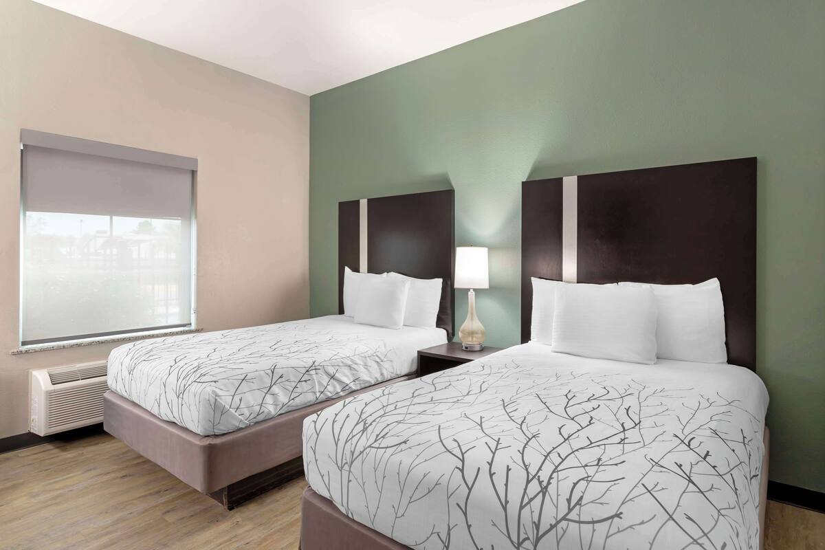 A hotel room with two beds, a window with blinds, a wall heater, and a nightstand with a lamp.