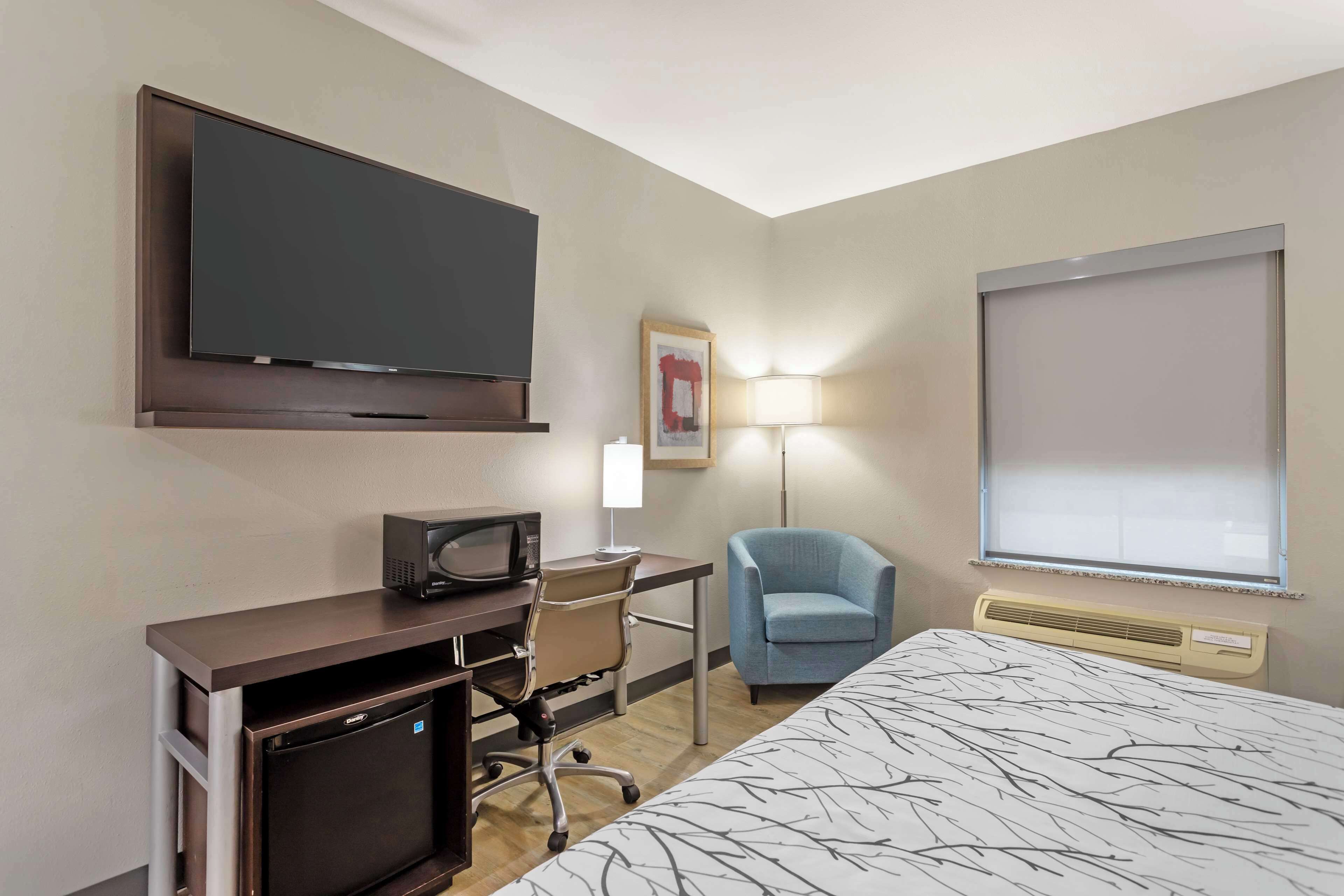 A hotel room with a bed, a desk with a computer, a television, a chair, a lamp, and a window.