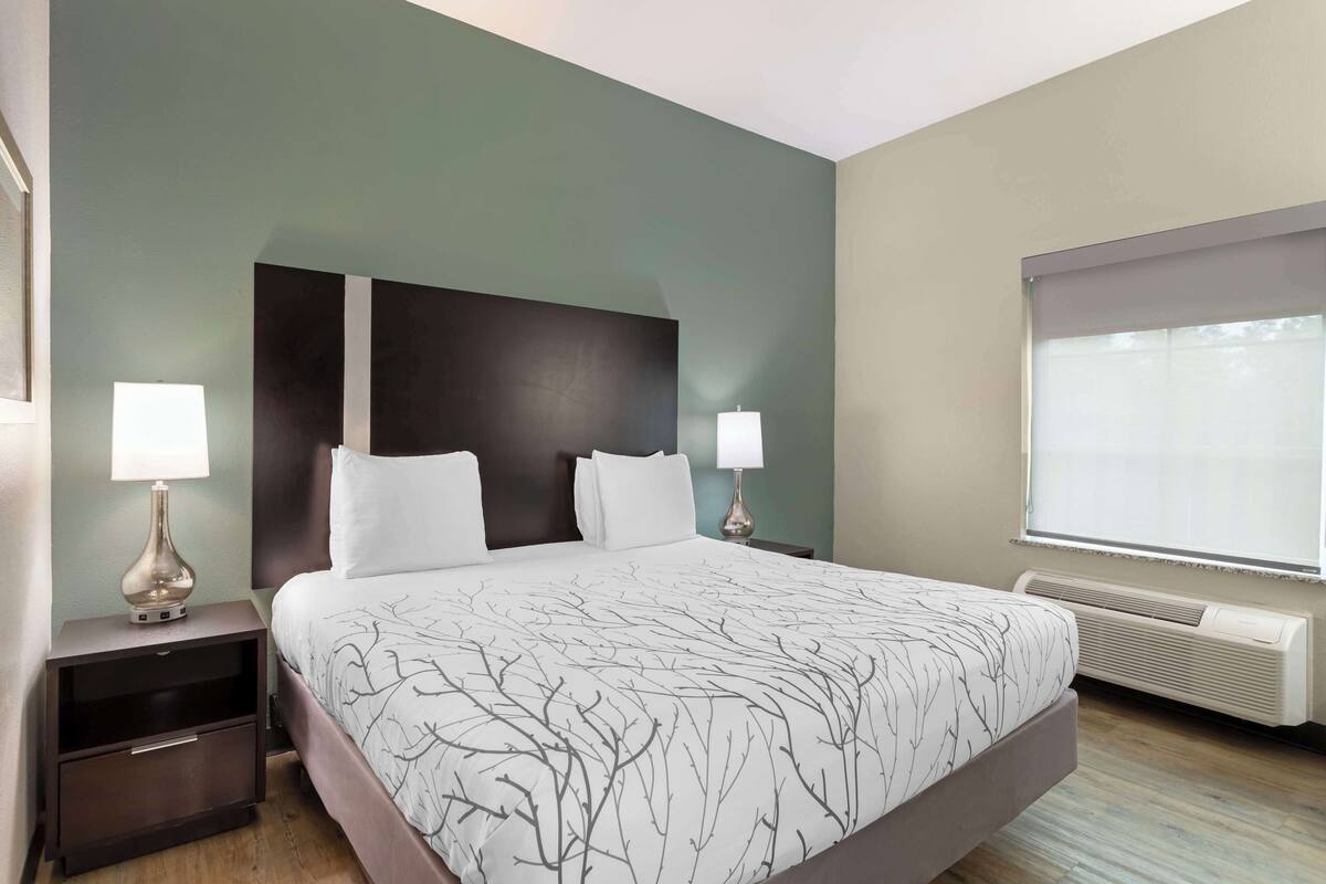 A hotel room with a large bed, two bedside tables with lamps, a window with blinds, and a wall-mounted air conditioner.