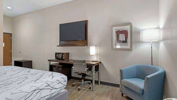 A hotel room with a bed, a desk with a computer, a TV, a chair, and a lamp.