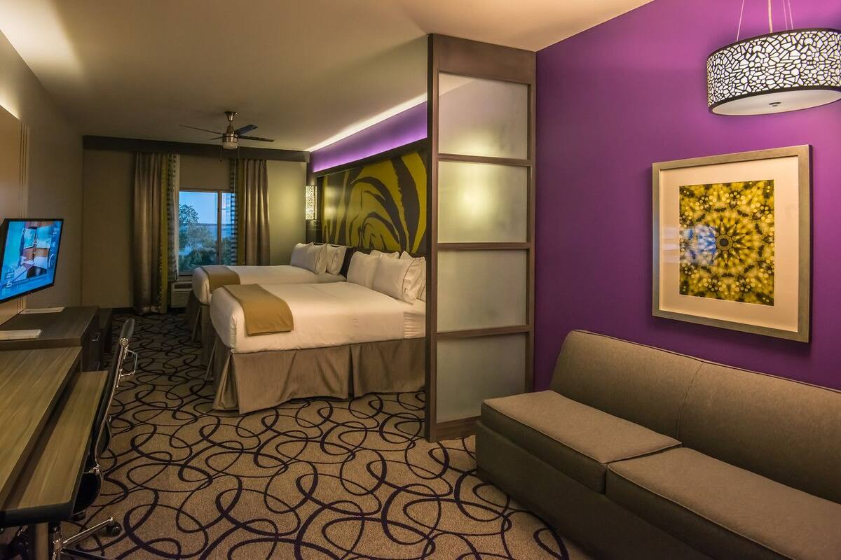 Ein Hotelzimmer mit einem Bett, einem Schreibtisch mit Telefon, einem Fernseher, einer Couch und einem Deckenventilator.