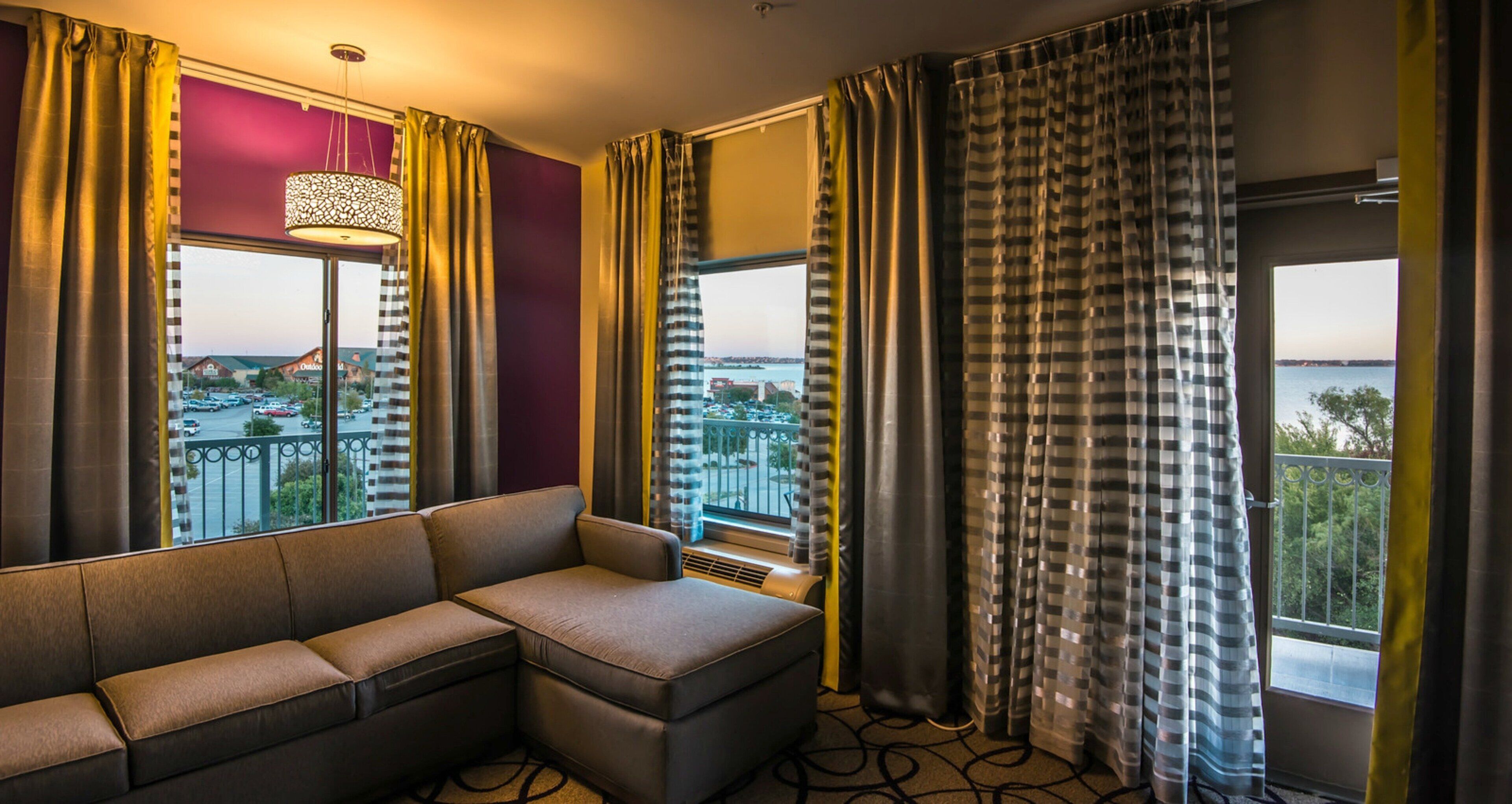 A hotel room with a sectional sofa, large windows with curtains, a hanging light fixture, and a view of a cityscape.