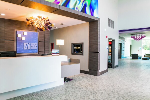 A hotel reception area with a modern design, including a reception desk, a digital display, and a seating area with a view of the hallway.