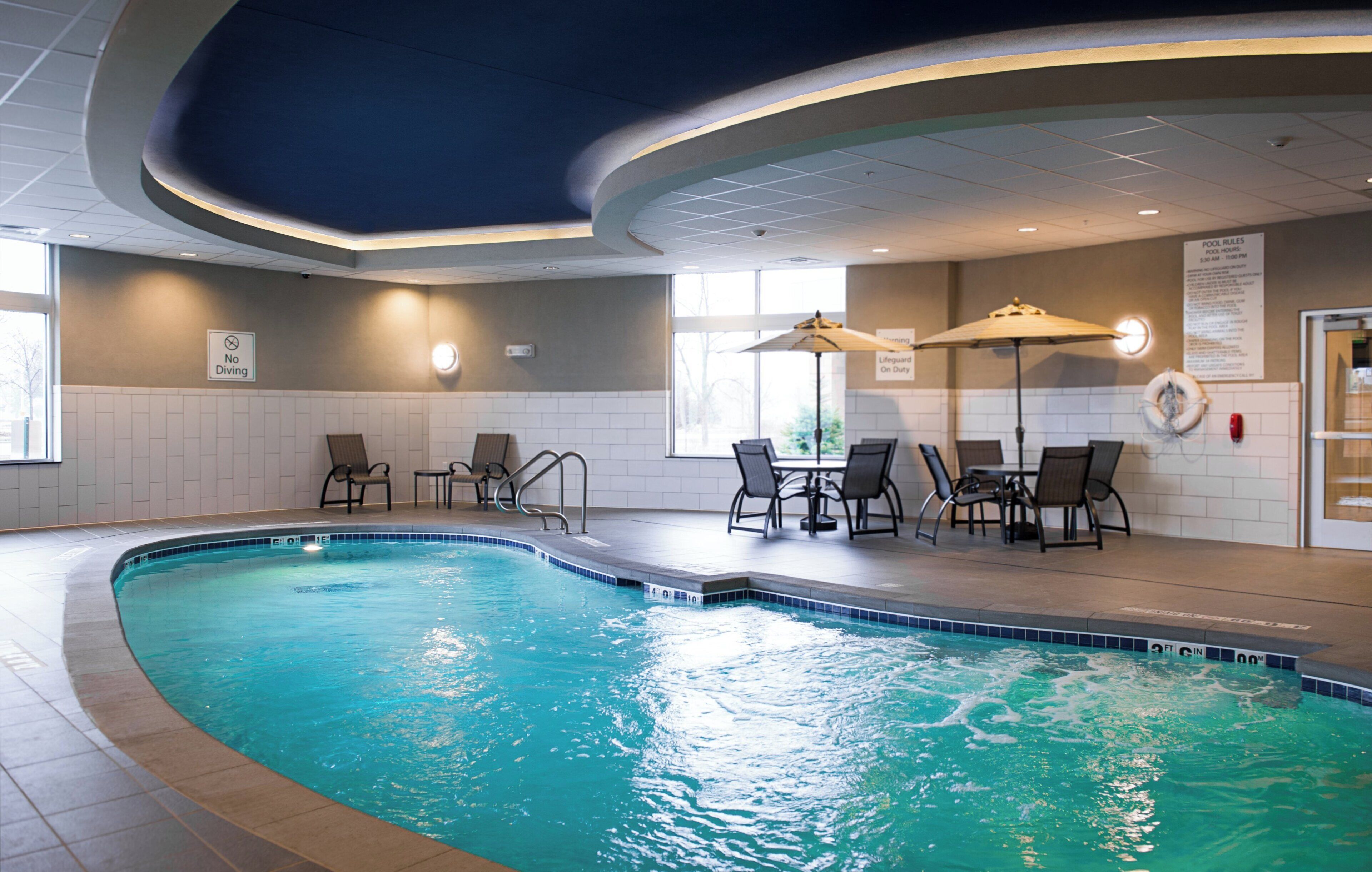 Una piscina interior con borde curvo, rodeada de sillones y mesas, y un cartel que indica la presencia de un socorrista.