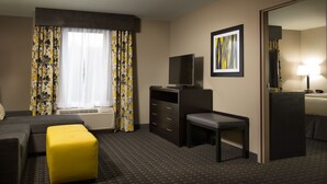 Une chambre d’hôtel avec un canapé, un pouf jaune, un bureau, une télévision, un tableau encadré et une lampe.