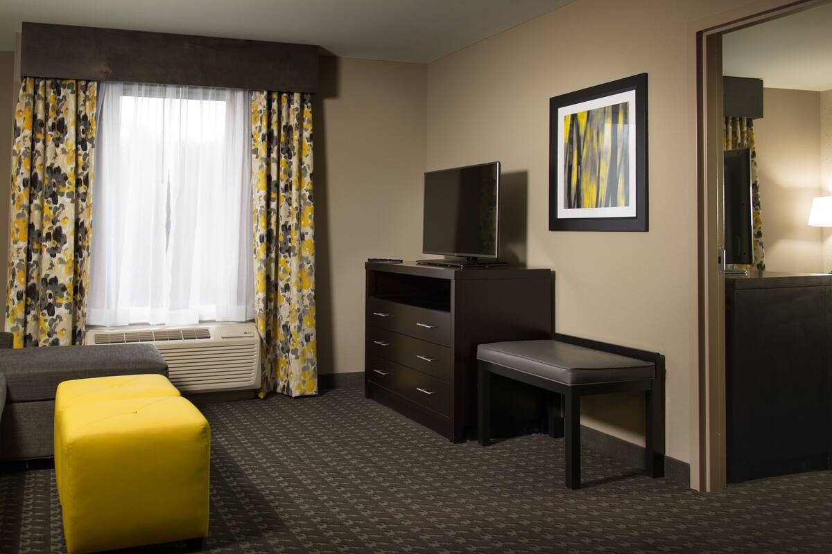 A hotel room with a sofa, a yellow ottoman, a desk, a television, a framed picture, and a lamp.