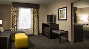 Quarto de hotel com sofá, um banquinho amarelo, escrivaninha, televisão, quadro emoldurado e abajur.
