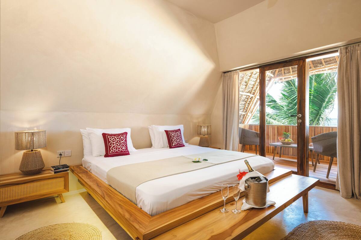 Habitación de hotel con una cama grande, cabecero de madera, mesita de noche con lámpara, un banco de madera y un balcón con vistas a palmeras.