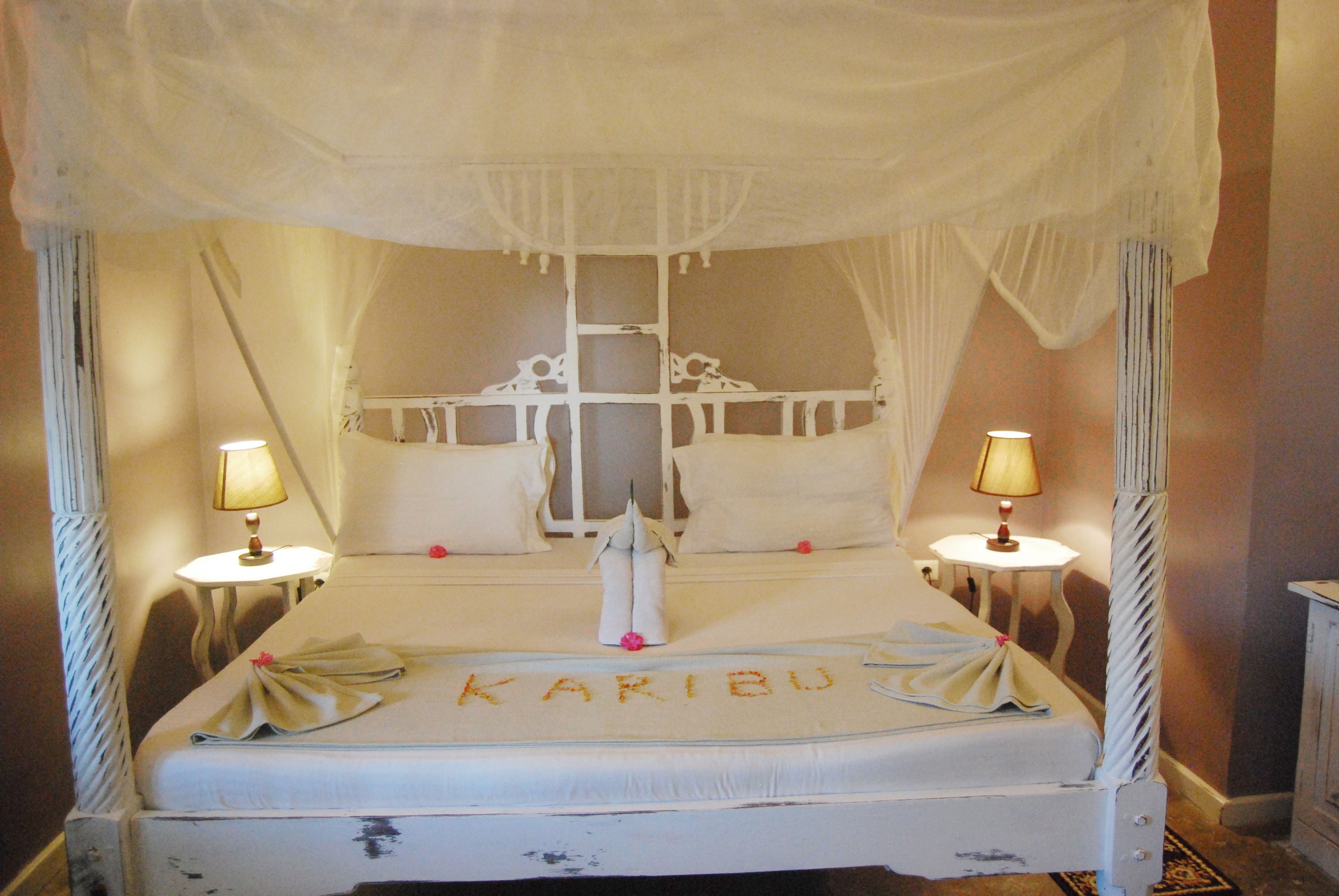 A four-poster canopy bed with a canopy, two bedside tables with lamps, and a bedspread with the name "KARIBU" embroidered on it.