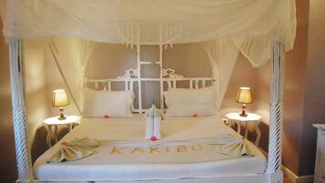 A four-poster canopy bed with a canopy, two bedside tables with lamps, and a bedspread with the name "KARIBU" embroidered on it.