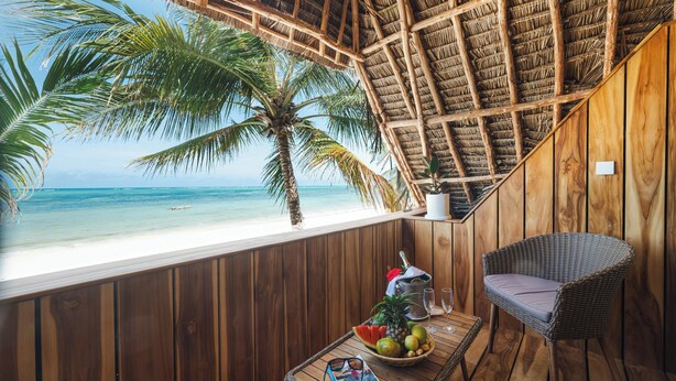 A wooden room with a view of the beach, a wicker chair, a table with fruits and glasses, and a book. - Sahari Zanzibar (Bwejuu)
