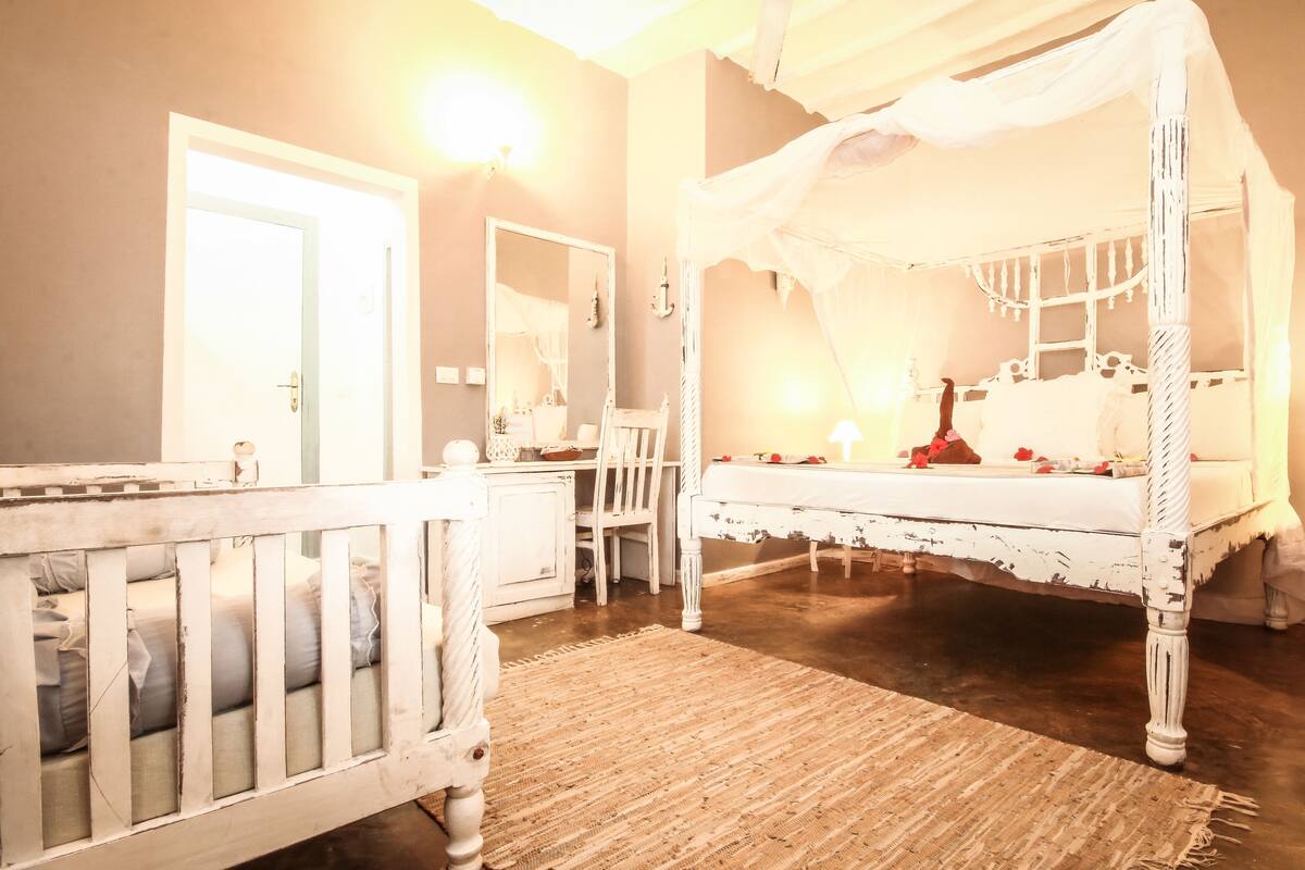 Una habitación con una cama con dosel, una cuna, un tocador con espejo y una puerta.