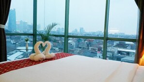 A hotel room with a large window, a bed with a red and white bedspread, and a heart-shaped decoration made of swans on the windowsill. - Business Hotel Tomang (Jakarta)