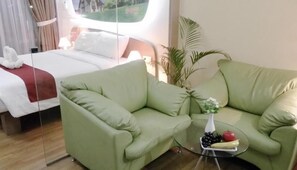 A hotel room with a green sofa, a glass coffee table with fruits, a bed with white bedding, and a view of greenery outside. - Business Hotel Tomang (Jakarta)