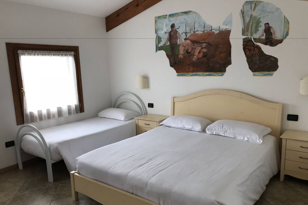 A bedroom with two single beds, a large bed, a window with curtains, and a wall with artwork.