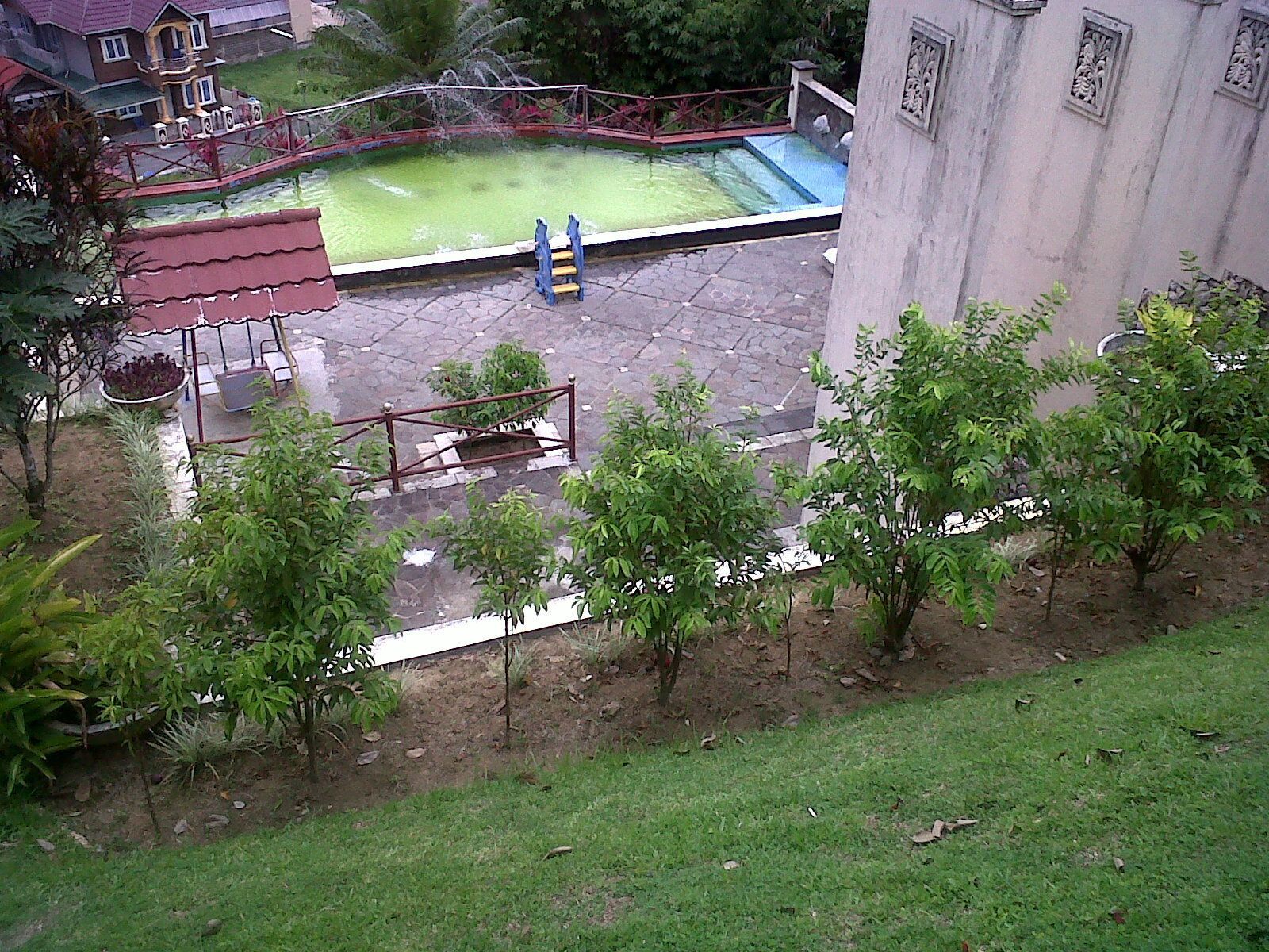 Una piscina amb una tanca vermella, una petita zona de jardí amb arbres i un edifici amb elements decoratius.