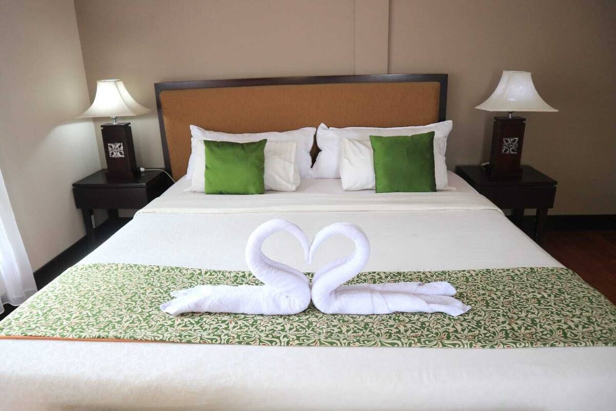 A neatly made bed with a green and white patterned bedspread, two white pillows, and a decorative swan towel arrangement.
