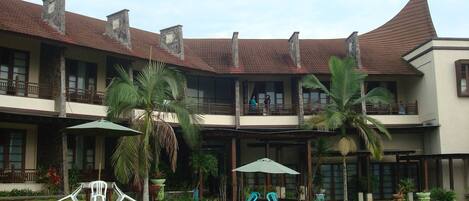 A two-story building with a curved roof, balconies, and a garden area with chairs and umbrellas.