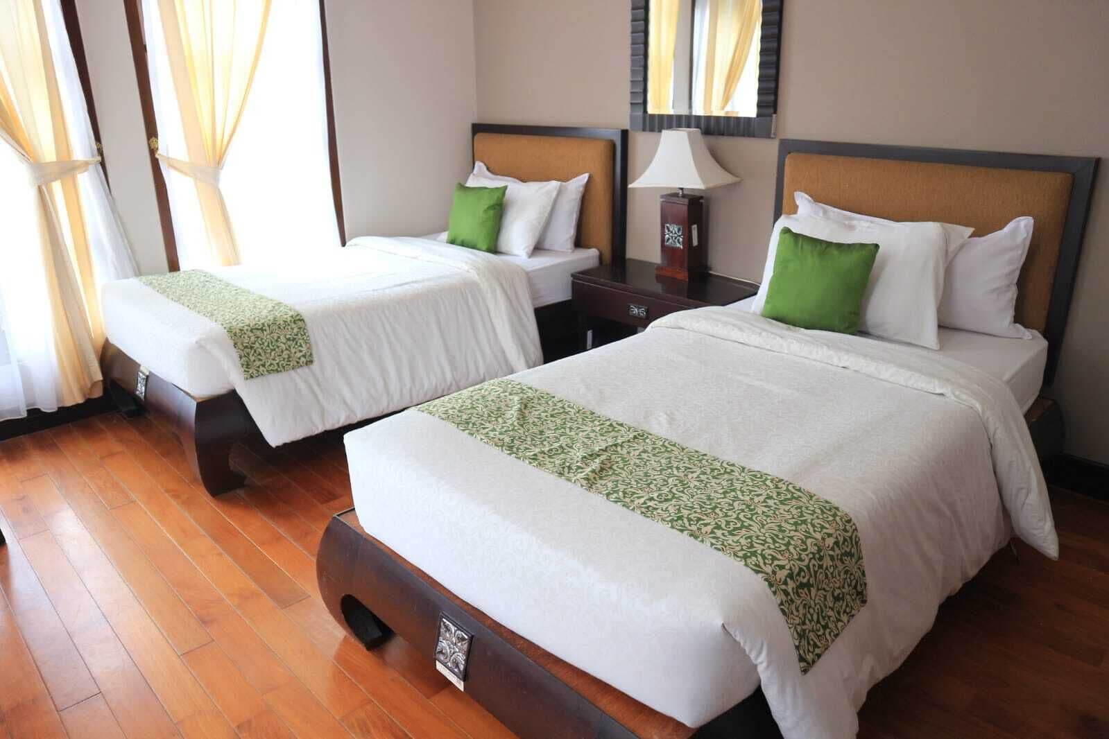 A hotel room with two beds, each with white bedding and green throw pillows. There is a nightstand with a lamp between the beds.
