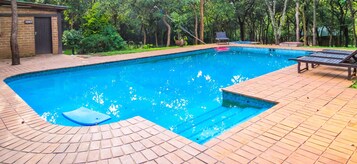 Una piscina rectangular con un borde de baldosas, rodeada de árboles y un pequeño edificio con una puerta que indica 'MEN'S'.