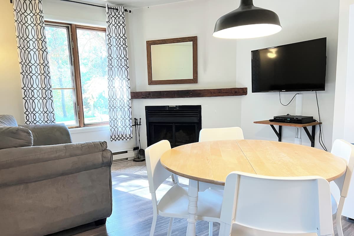 A living room with a sofa, a dining table with chairs, a fireplace, and a TV mounted on the wall.