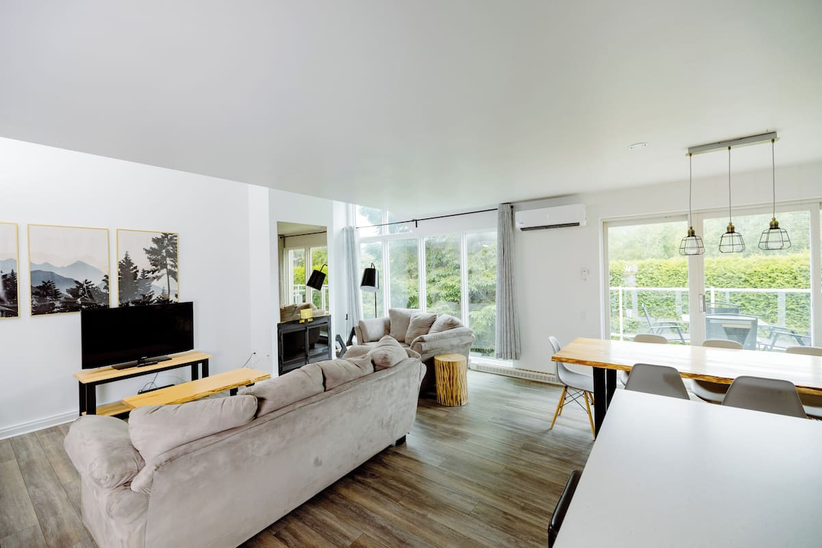 A modern living room with a sofa, TV, dining table, and large windows.