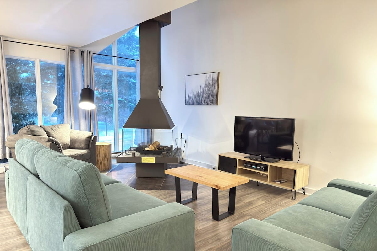 A modern living room with a sofa set, a TV, a fireplace, and large windows.