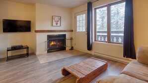 Una sala de estar con chimenea, televisor montado en la pared y ventana con vistas a árboles.