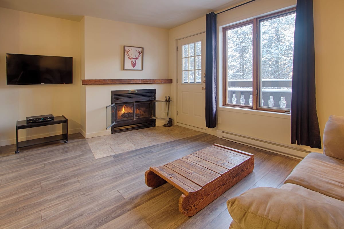 A living room with a fireplace, a television mounted on the wall, and a window with a view of trees.