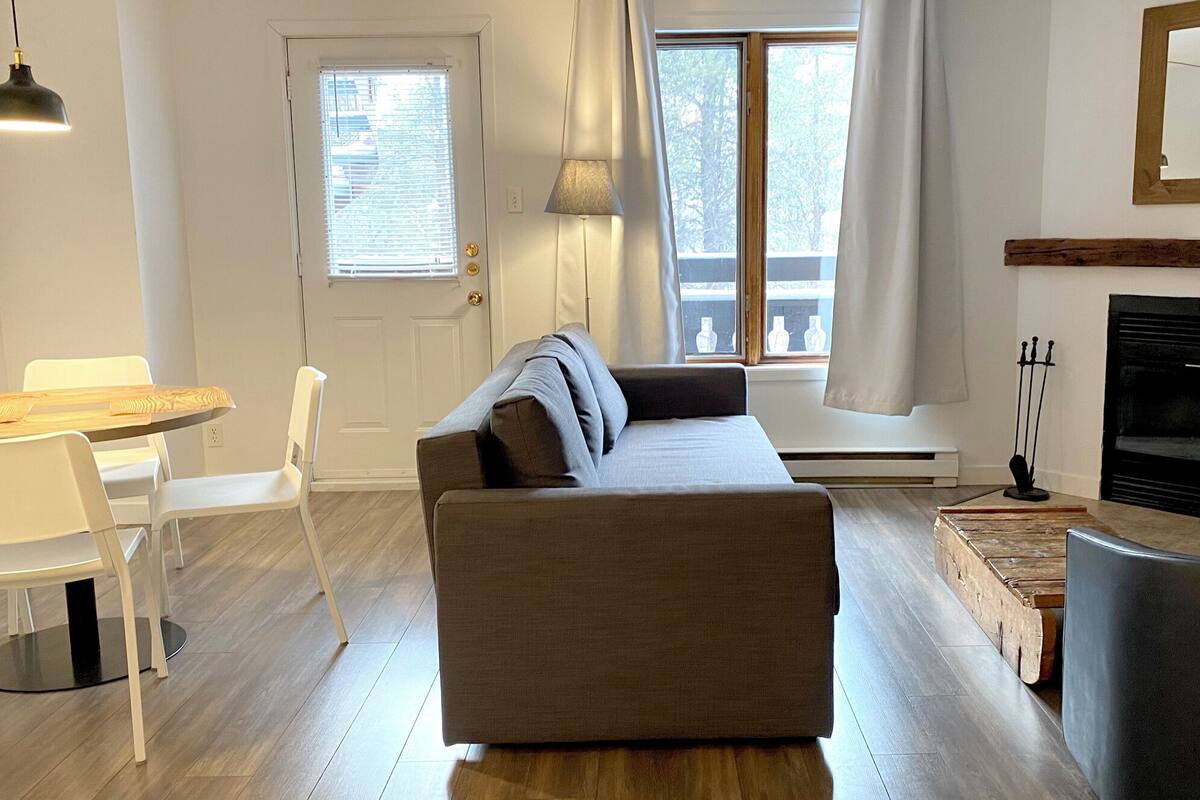A modern living room with a sofa, a dining table with chairs, a fireplace, and a large window with blinds.