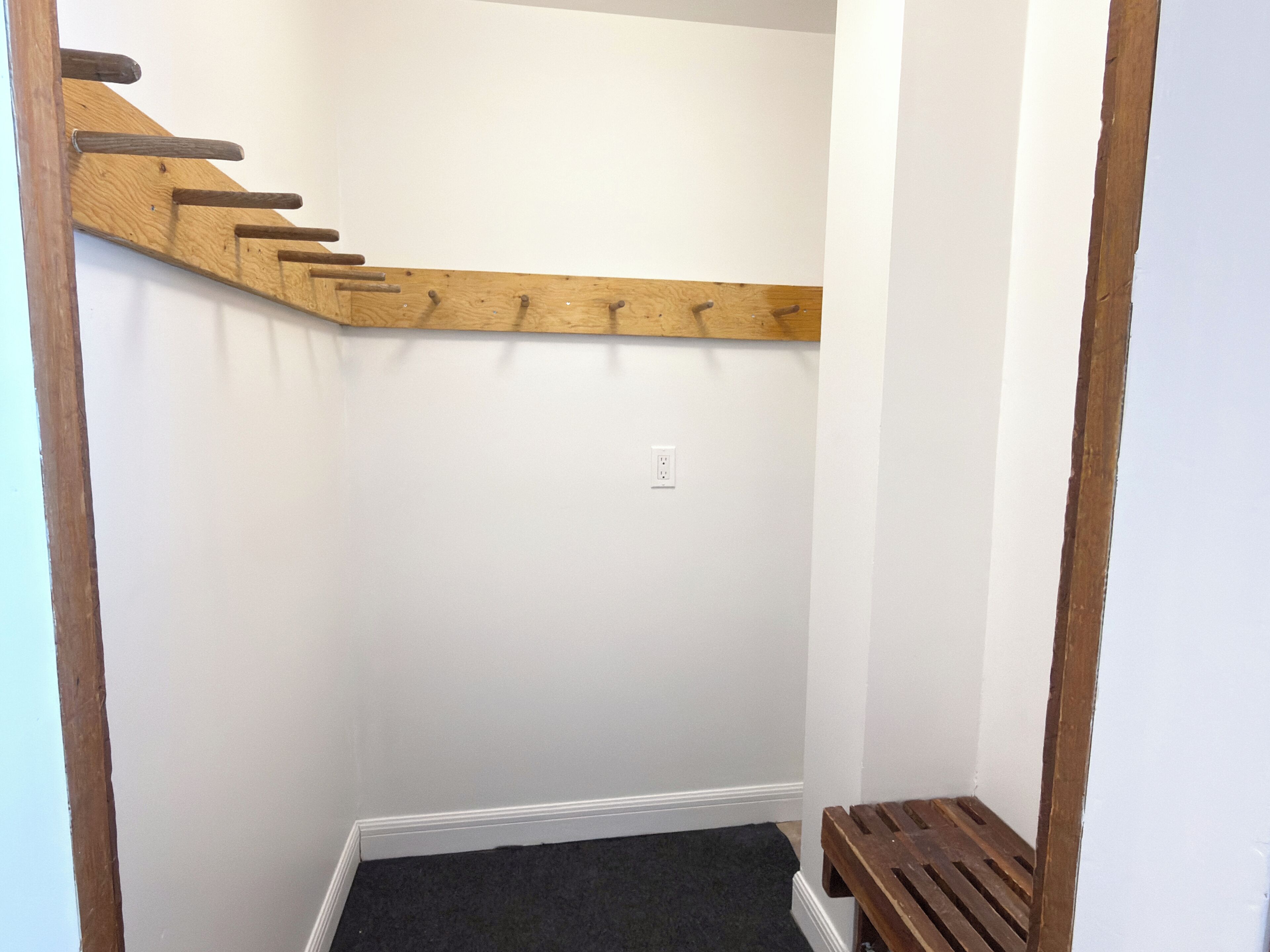 A corner of a room with a wooden bench, a white wall, and a black carpet.