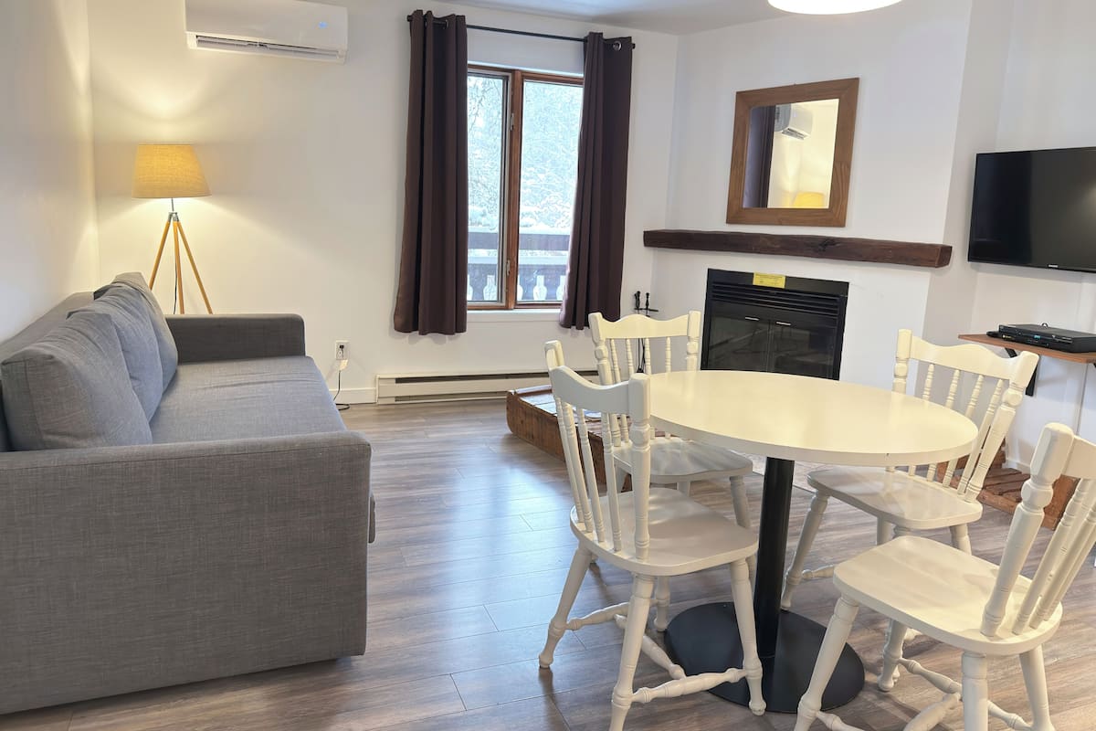 A modern living room with a grey sofa, white chairs, a round table, a fireplace, and a TV mounted on the wall.