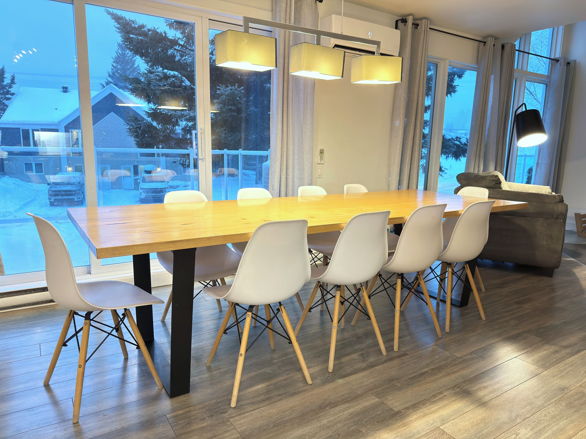 A modern dining area with a wooden table and white chairs, large windows with a view of a snowy landscape, and a grey sofa.