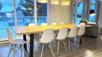 A modern dining area with a wooden table and white chairs, large windows with a view of a snowy landscape, and a grey sofa.