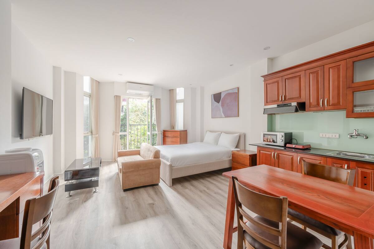 A modern hotel room with a bed, a sofa, a dining table, a kitchen area, and a TV.