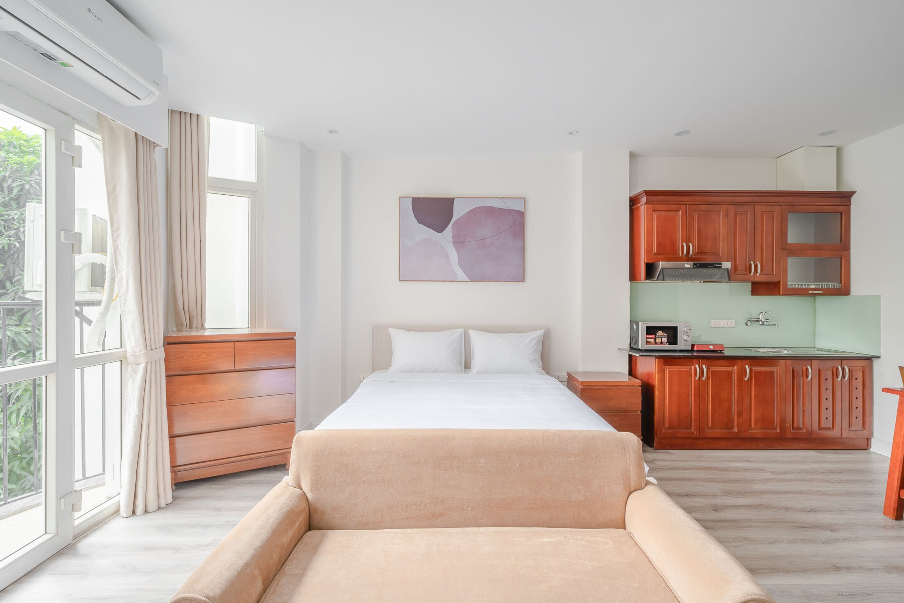 A bedroom with a bed, a sofa, a window with curtains, a kitchen area with cabinets, and a wall-mounted artwork.