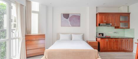 A bedroom with a bed, a sofa, a window with curtains, a kitchen area with cabinets, and a wall-mounted artwork.