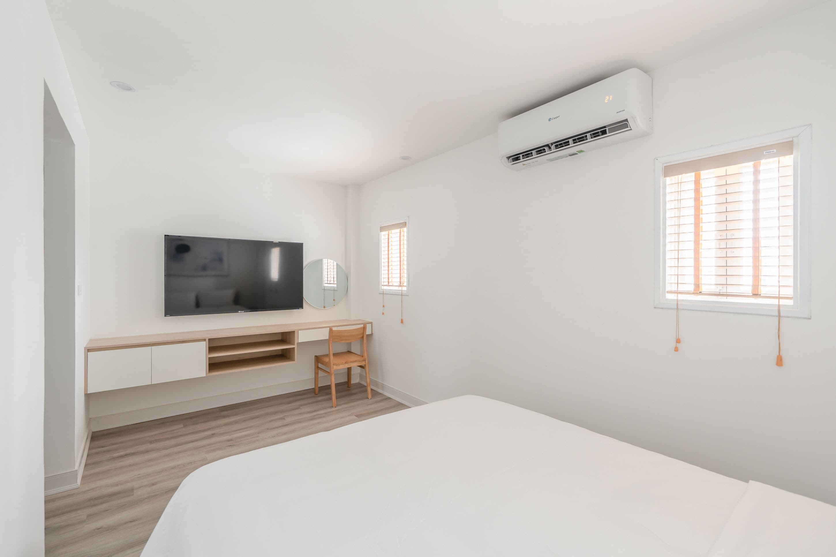 A modern bedroom with a bed, a desk with a chair, a TV, and two windows with blinds.