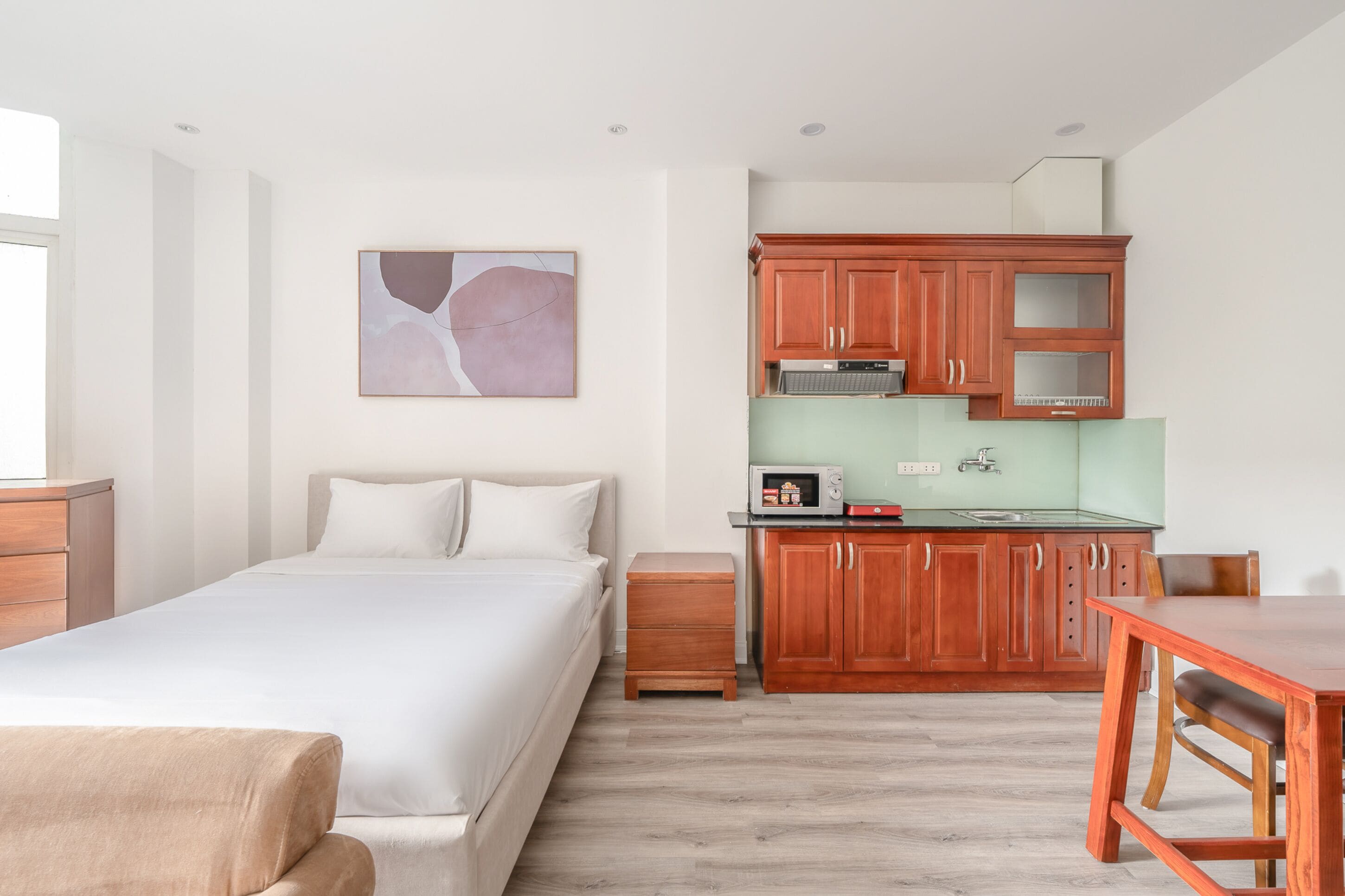 A bedroom with a bed, a small wooden nightstand, a kitchenette with wooden cabinets and a microwave, and a painting on the wall.