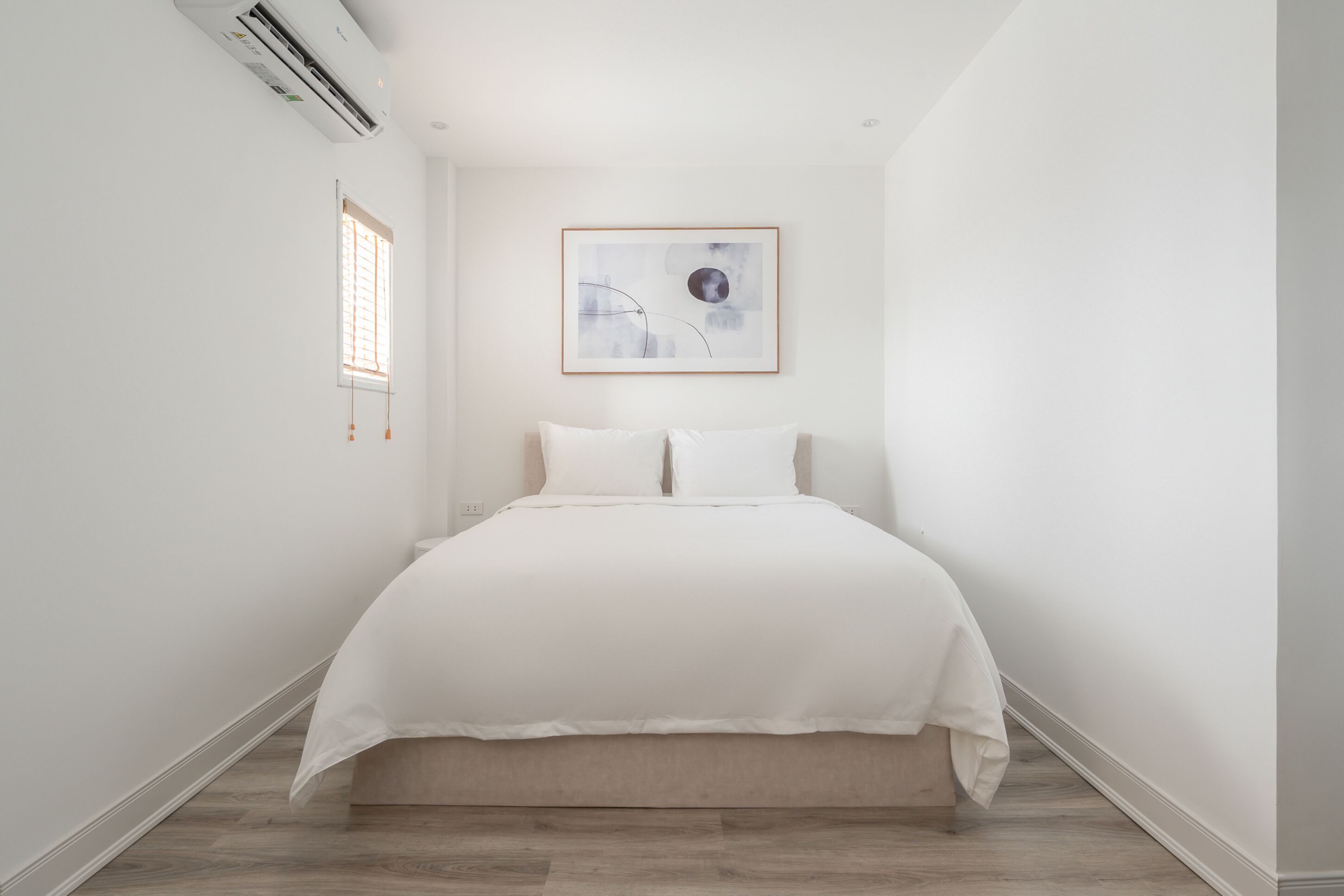 A minimalist bedroom with a large bed, a framed abstract painting, and a window with blinds.